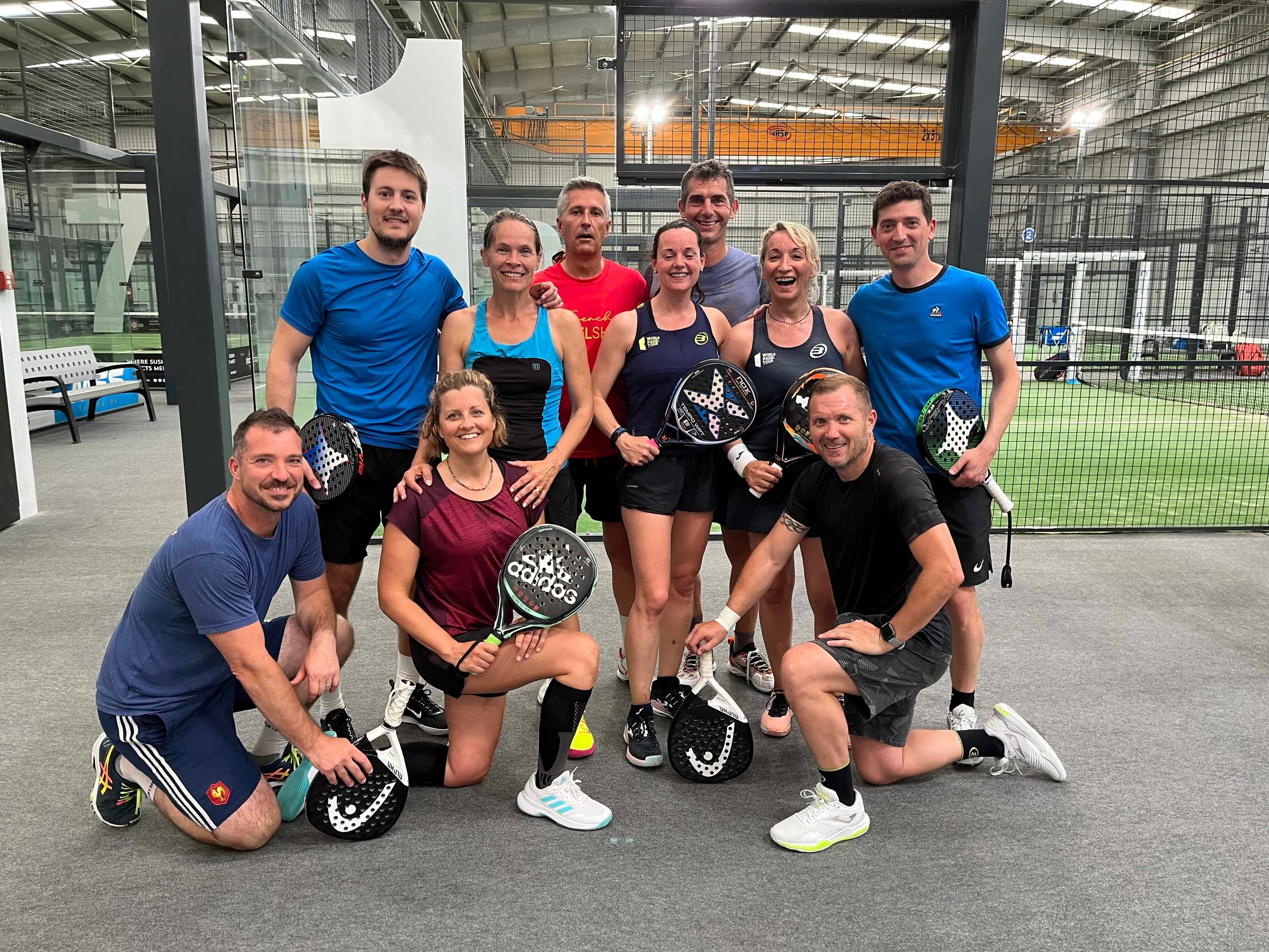 A group of nine people posing with padel rackets inside an indoor sports facility, with courts visible in the background.