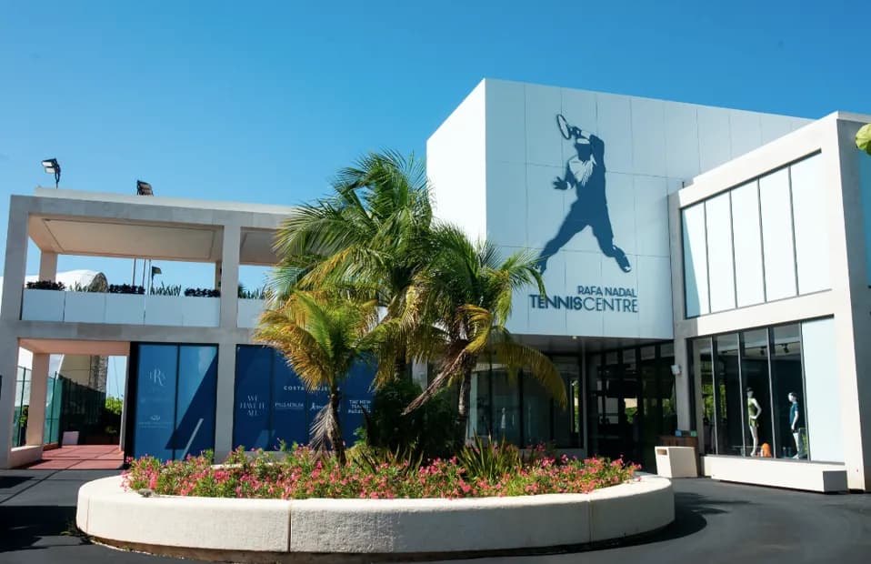 Exterior view of the International Tennis Centre in Cancún, Mexico, featuring a modern white facade with large glass windows, a sign with a tennis player silhouette, and palm trees under a clear blue sky.