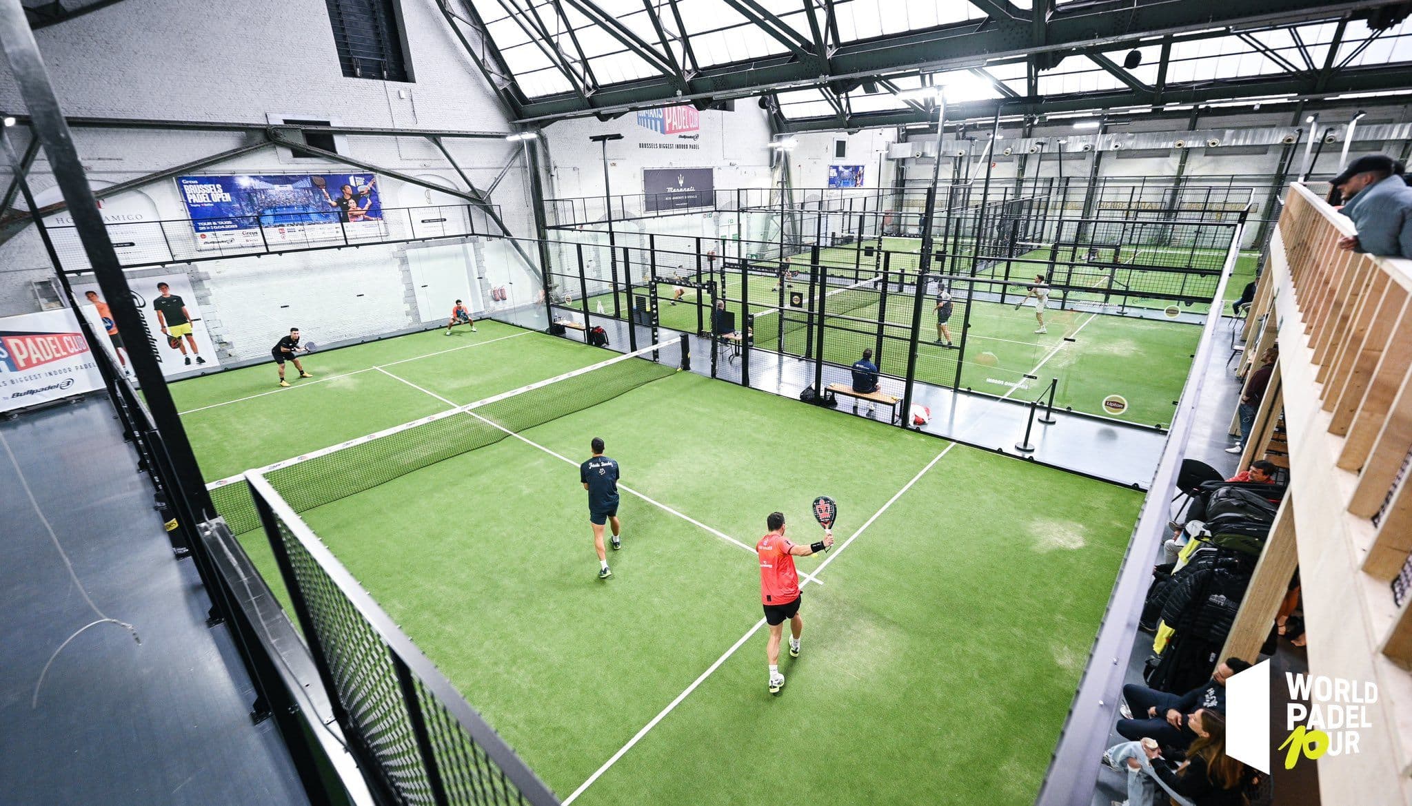 Indoor padel court with players, glass walls, and seating area.