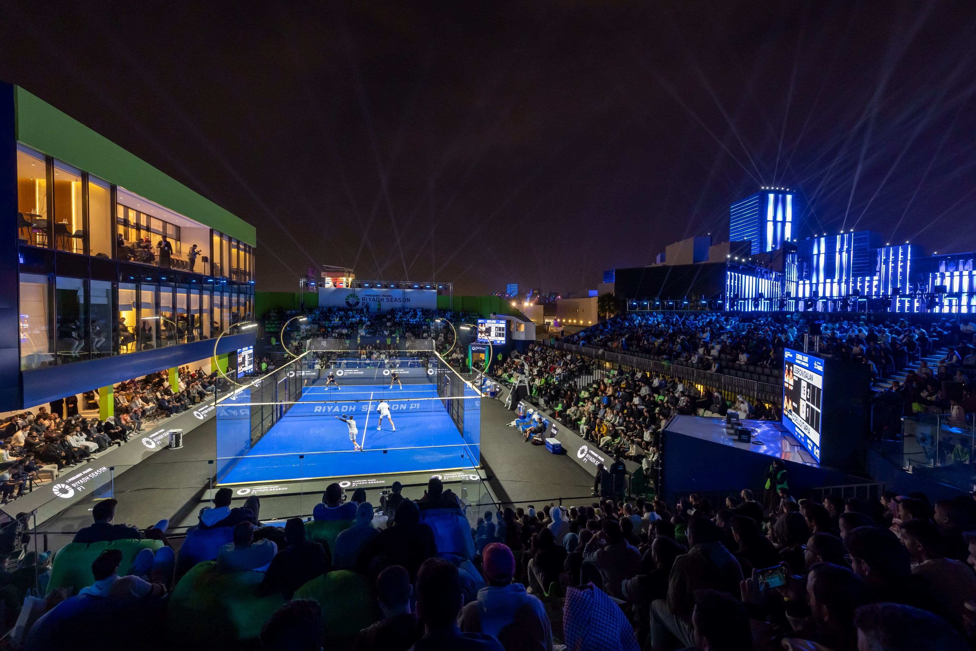 Nighttime padel match in a stadium with bright lights and a lively crowd.