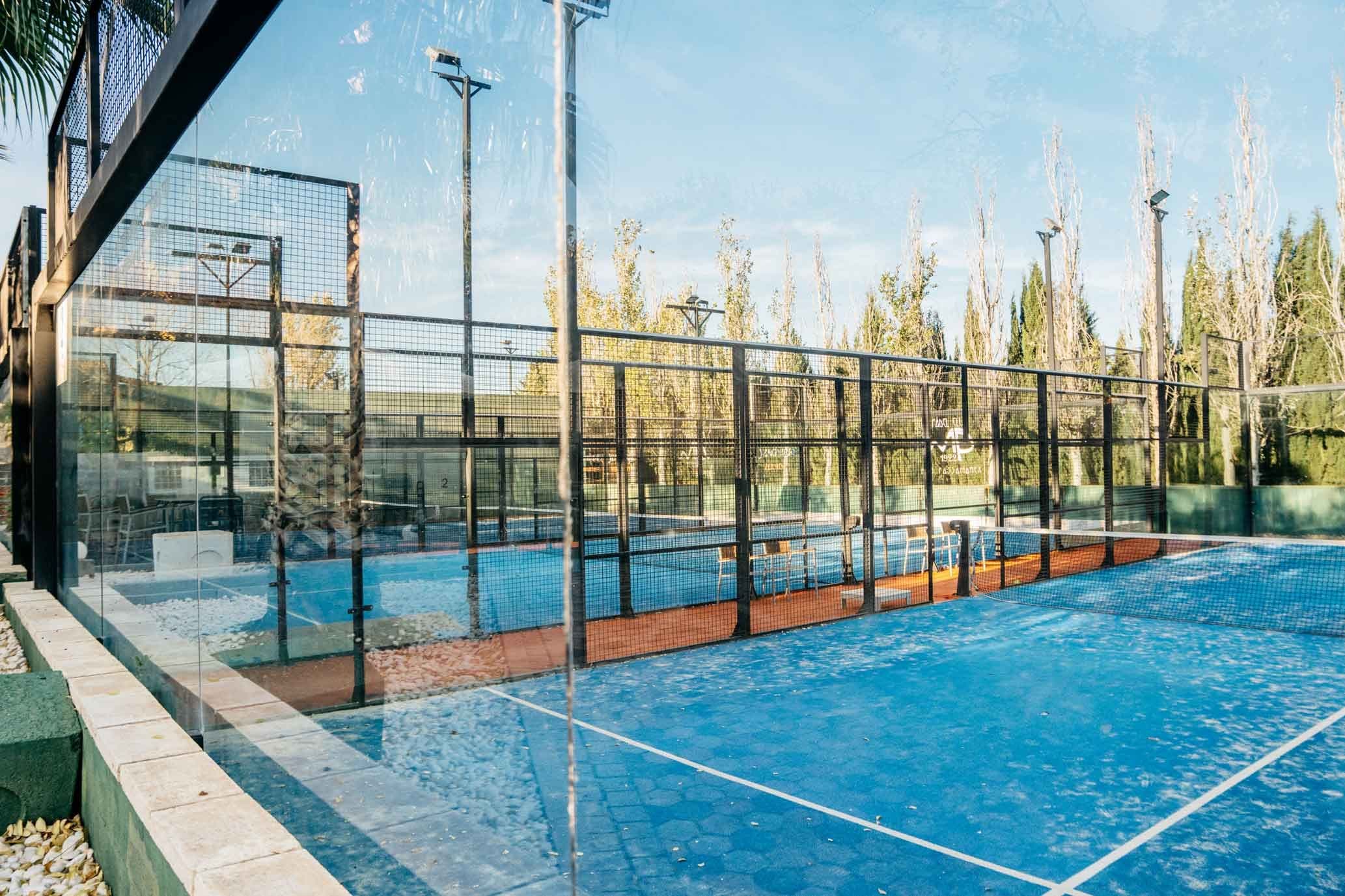 An outdoor padel court at Boutique Hotel Mas Passamaner, featuring a blue playing surface enclosed by glass walls and metal fencing, surrounded by greenery and tall light poles for evening matches.