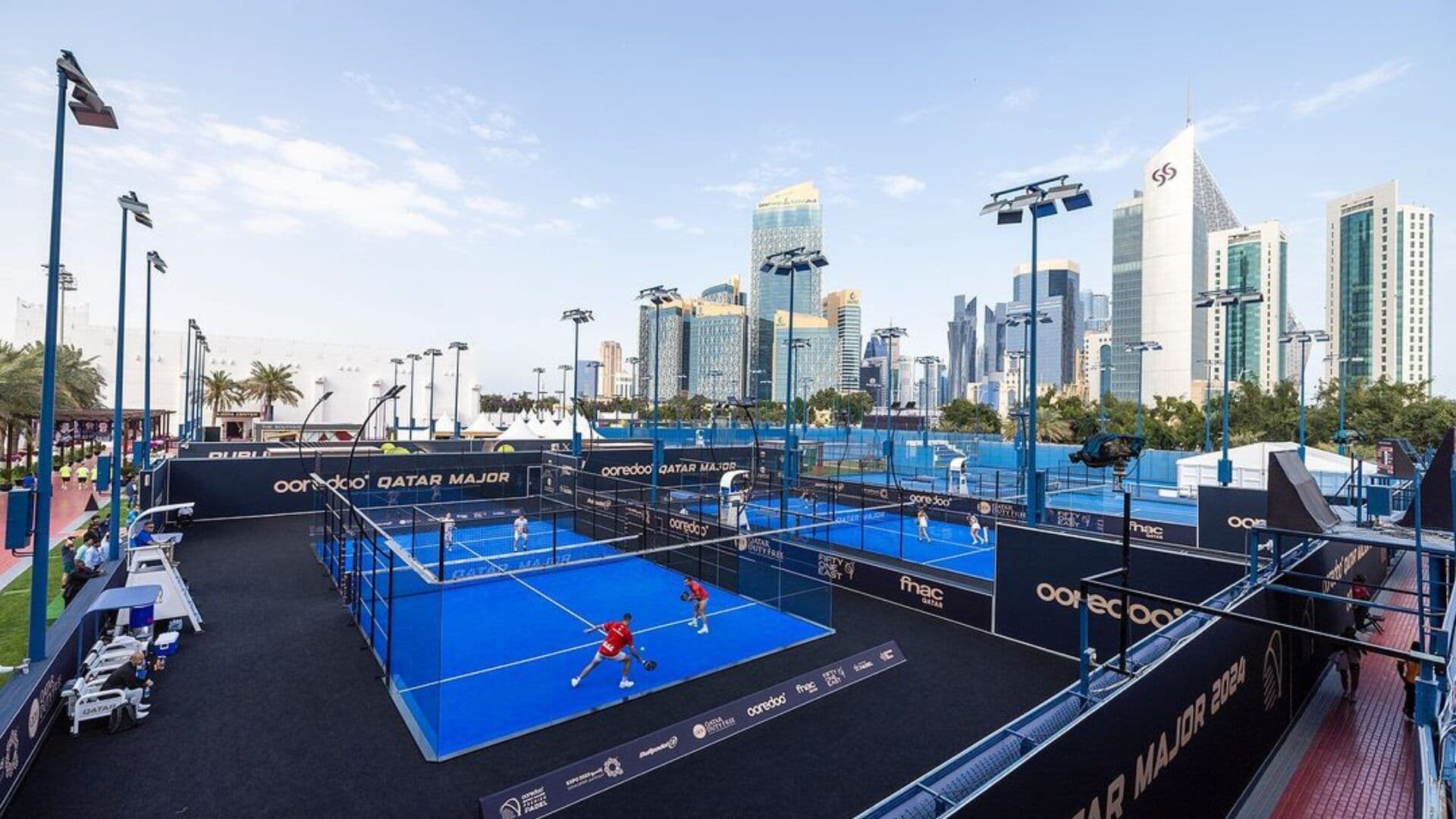 Outdoor padel courts at the Qatar Major Premier Padel event, featuring blue playing surfaces surrounded by black fencing, equipped with lighting for evening matches, set against a backdrop of modern skyscrapers in an urban environment.