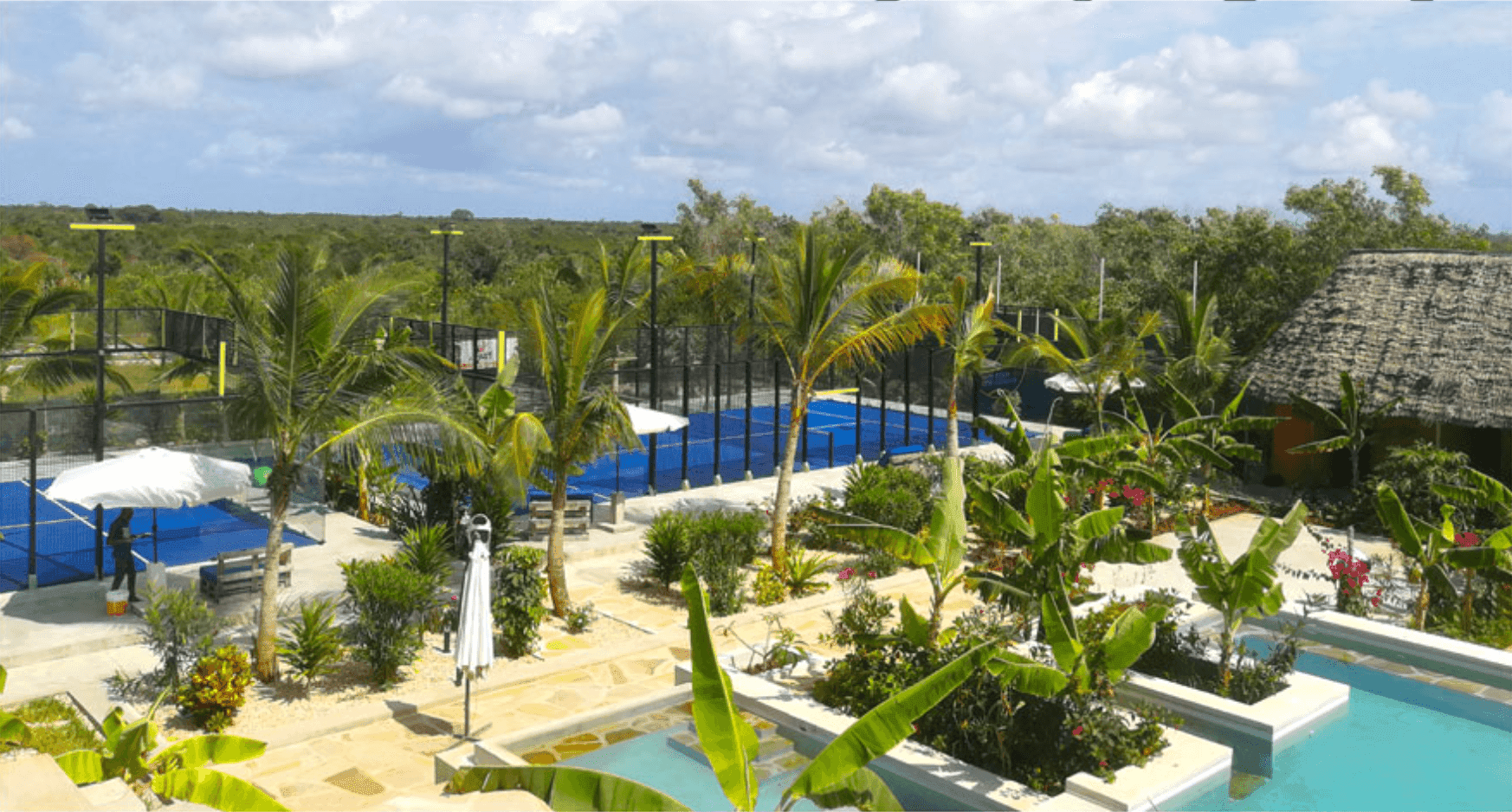 A serene view of Zanzibar Sporting Residence & Club featuring a swimming pool surrounded by lush greenery, tennis courts, and palm trees under a partly cloudy sky.