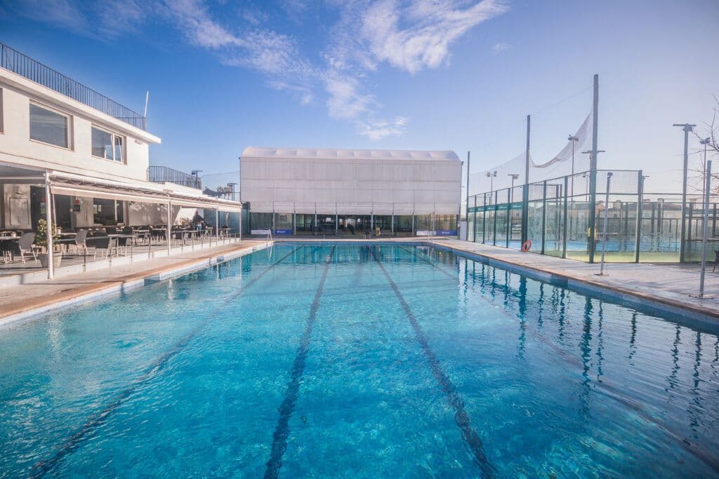 An outdoor swimming pool at Udyr Sport Club Mallorca, featuring clear blue water with lane markings, surrounded by sports facilities, including tennis or padel courts, and a building with seating areas under a clear sky.