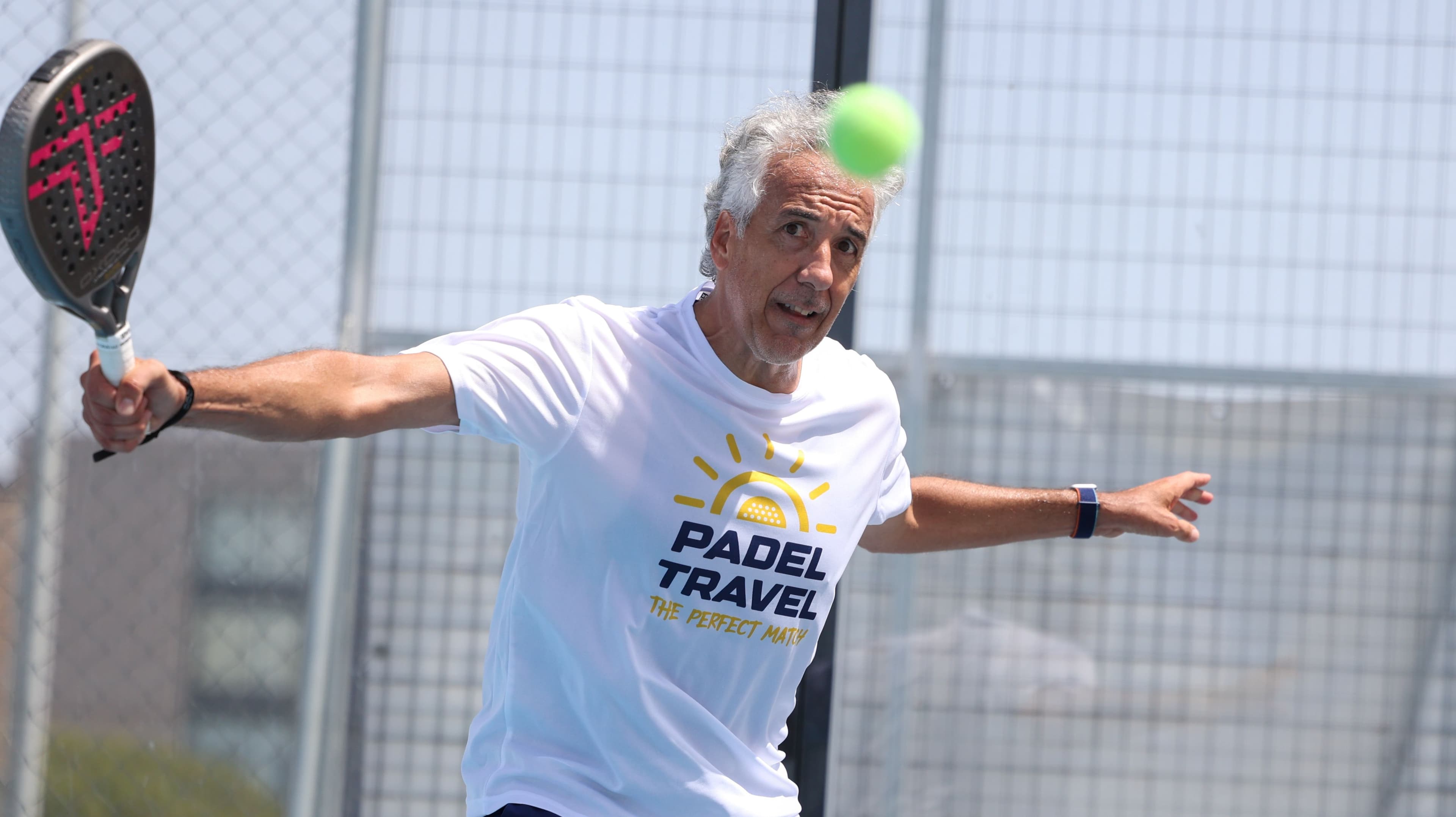 A padel player in mid-swing wearing a white t-shirt with "PADEL TRAVEL" printed on it, on a fenced court with buildings in the background.