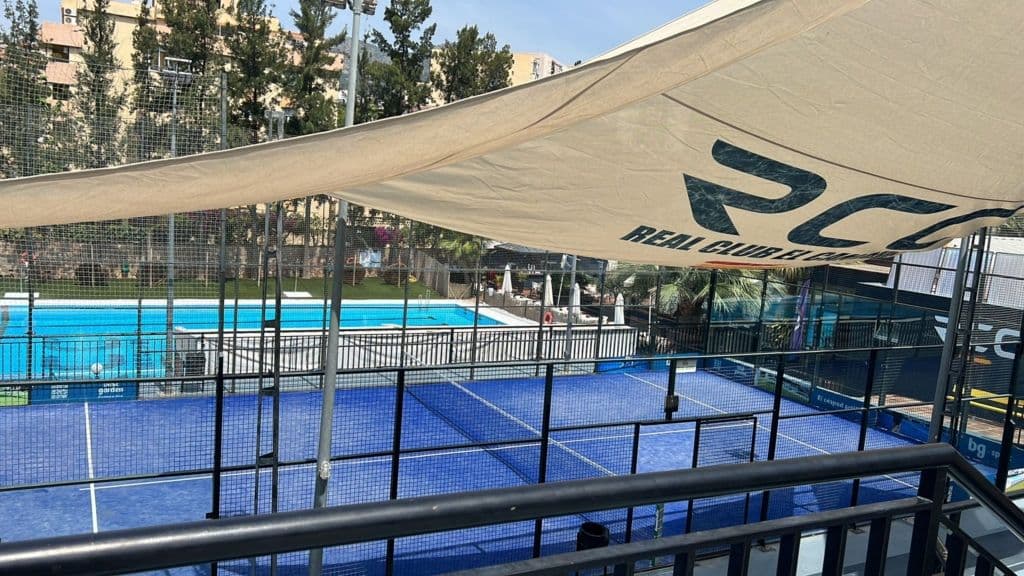 Outdoor padel court at Club Padel El Candado in Málaga, featuring a blue playing surface enclosed by glass walls, a large canopy with "RC Real Club El Candado" printed on it, and a swimming pool surrounded by trees in the background.