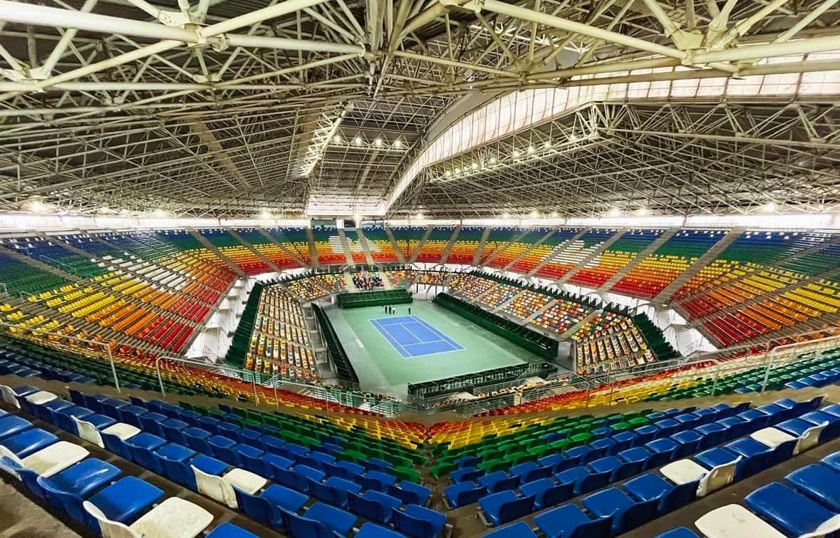 A large indoor sports arena in Buenos Aires filled with spectators, featuring a blue padel court at the center surrounded by glass walls, bright lights, and large screens displaying match details.