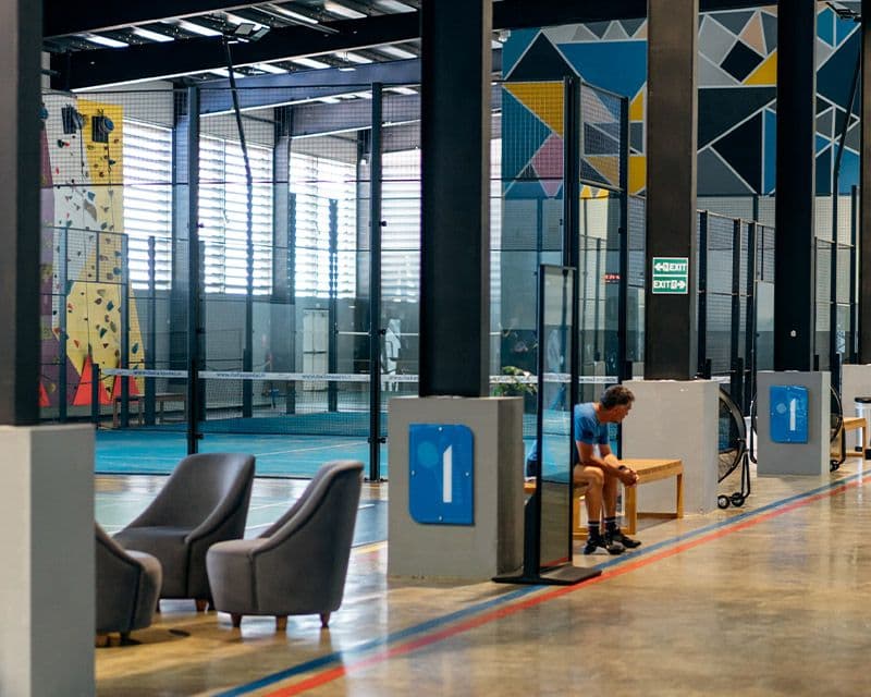 A modern indoor recreational area at RM Grand Baie featuring seating arrangements, a climbing wall, colorful floor patterns, and glass partitions under natural light streaming through large windows.